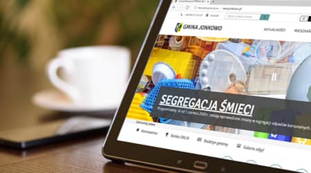 Źródło: Pixabay Tablet oparty na stole z wyświetloną stroną internetową gminy Jonkowo. W tle stojąca filiżanka.