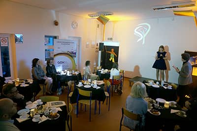 Zdjęcie przedstawiające osoby siedzące przy stolikach. W głębi osoba stojąca na scenie, mówiąca do mikrofonu.