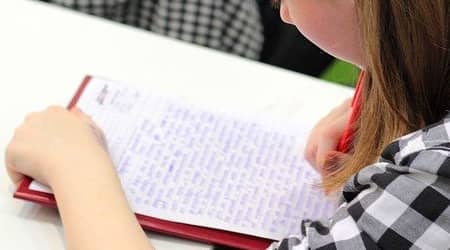 Zdjęcie wykonane od tyłu, przedstawiające młodą osobę piszącą długopisem na kartce długi tekst.