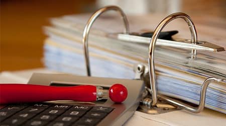 Zdjęcie przedstawiające otwarty segregator z dokumentami oraz długopis leżący na kalkulatorze.