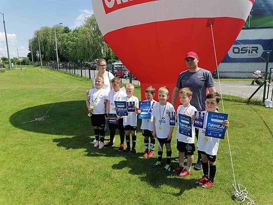 Zdjęcie przedszkolaków z trenerem i wychowawcą na tle banneru sponsorskiego.