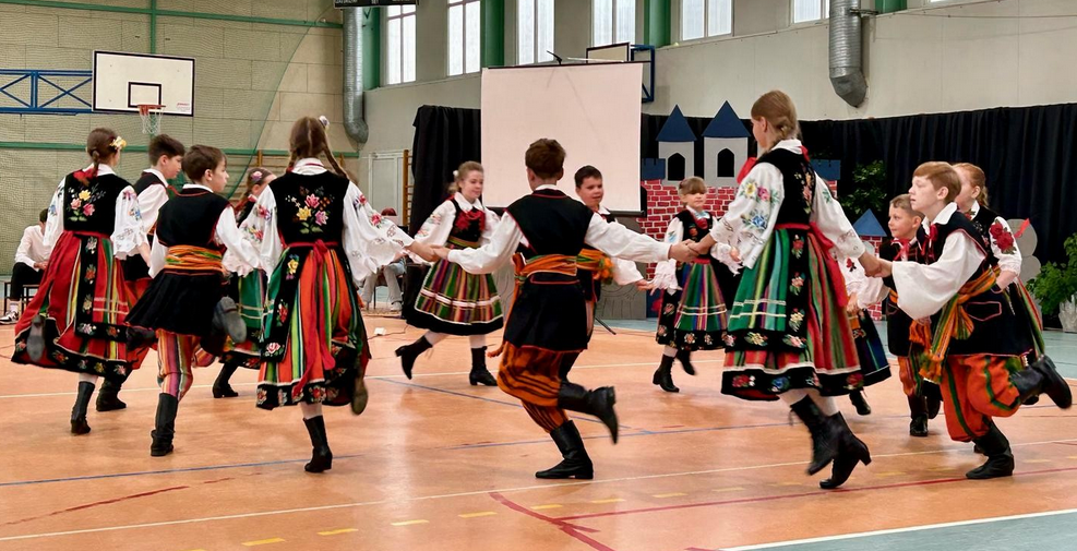 źródło: własne Na zdjęciu widać grupę młodych ludzi w tradycyjnych strojach ludowych, którzy wykonują taniec na sali gimnastycznej. Stroje są kolorowe, zdobione haftami, a dominują w nich barwy czerwieni, zieleni i czerni. Chłopcy mają na sobie białe koszule, czarne spodnie i haftowane kamizelki, a dziewczynki białe bluzki, haftowane gorsety i szerokie, wzorzyste spódnice. Tancerze trzymają się za ręce, tworząc okręgi lub inne figury taneczne, a ich pozy sugerują dynamiczny ruch. W tle widoczny jest parkiet sali gimnastycznej z namalowanymi liniami oraz elementy wyposażenia, takie jak tablica do koszykówki.